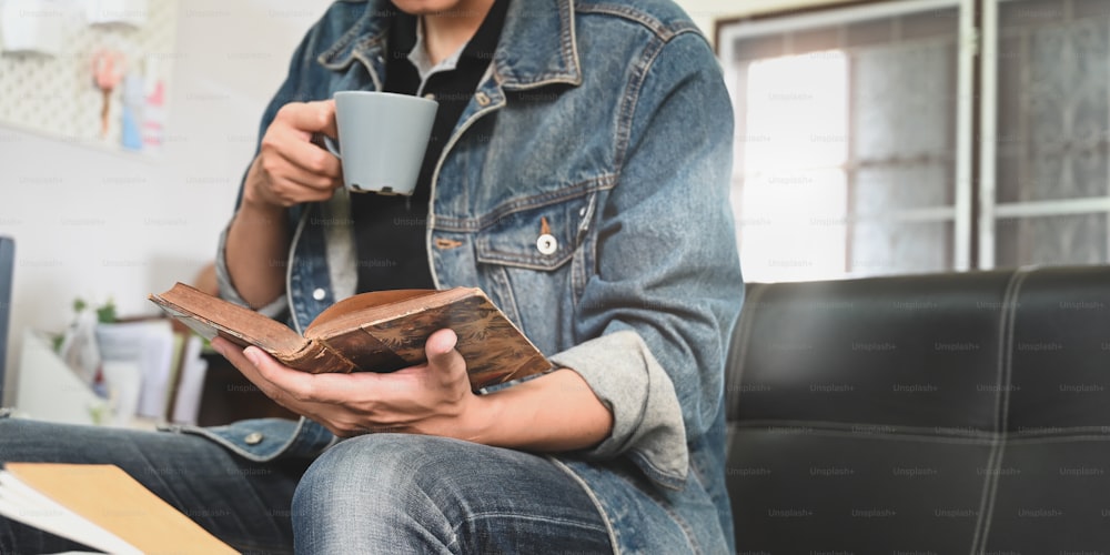 Cropped image of smart man drinking a hot coffee and holding/reading a book in his hand while sitting and relaxing at the black leather sofa over comfortable sitting room as background.