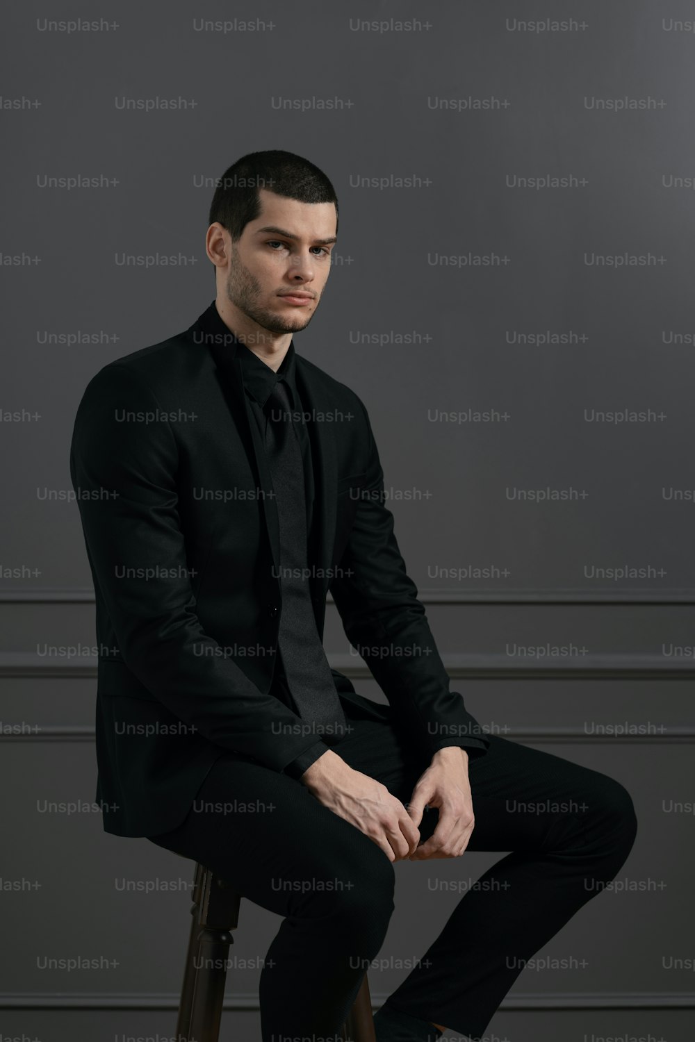 Junger gutaussehender Geschäftsmann in schwarzem Hemd und schwarzem Anzug. Selbstbewusster junger Geschäftsmann posiert auf grauem Hintergrund auf Stuhl sitzend