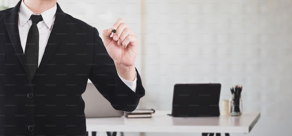 Imagen recortada de hombre de negocios en traje formal negro apuntando el bolígrafo para escribir en el tablero mientras está de pie sobre el escritorio de trabajo rodeado de oficina.