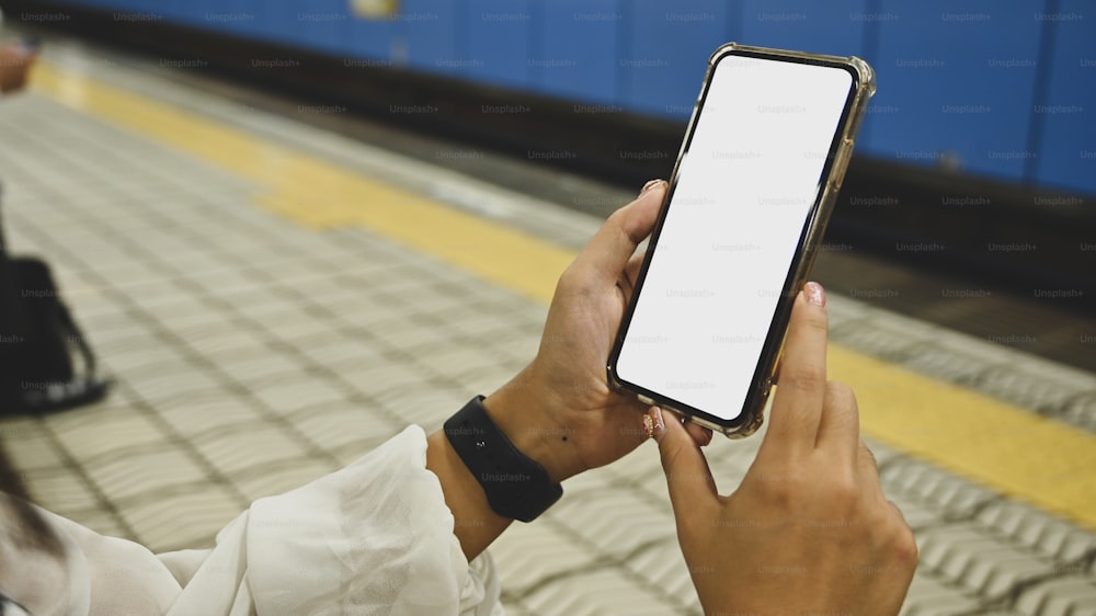Immagine ritagliata di mani di donna attraente che tengono uno smartphone nero con schermo bianco vuoto su una piattaforma ferroviaria vuota come sfondo.