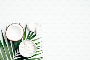 Frutto di cocco con metà e cocco grattugiato in ciotola, foglia di palma tropicale su sfondo bianco. Posa piatta, vista dall'alto. Sfondo estivo.