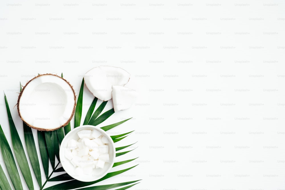 Fruit de noix de coco avec demi-noix de coco et noix de coco râpée dans un bol, feuille de palmier tropical sur fond blanc. Pose à plat, vue de dessus. Fond d’été.