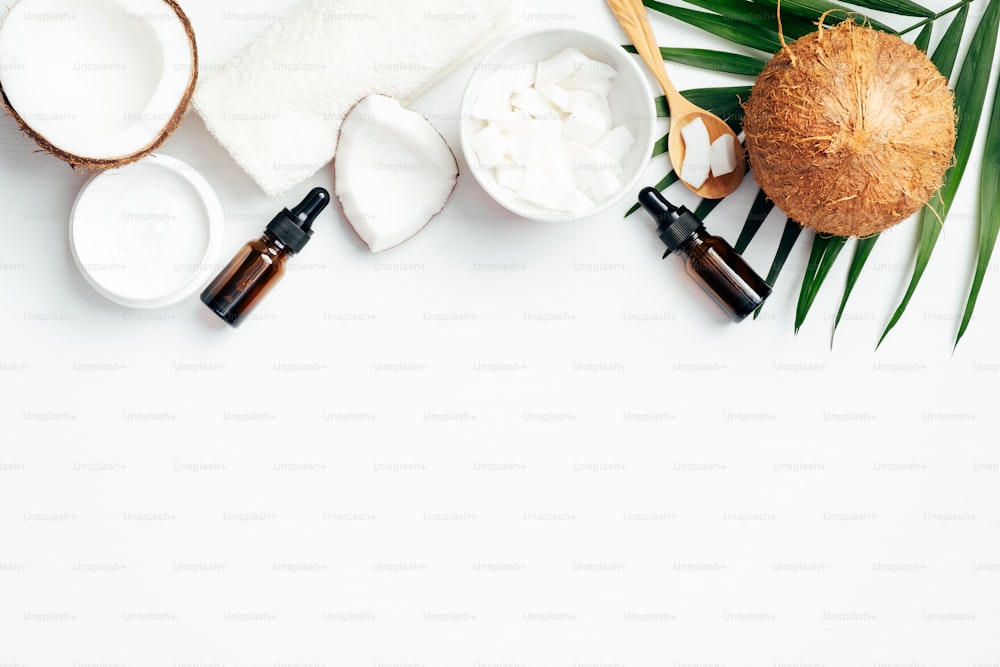 Set of natural coconut cosmetic on white background. Flat lay composition with coconut oil, hand cream, coconut half, grated coconut in bowl, tropical palm leaf and towel. SPA organic beauty products