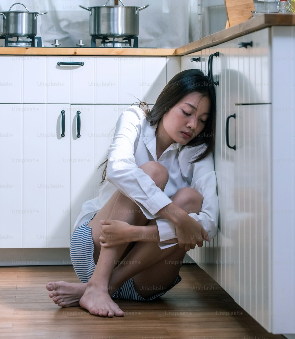 Tristeza hermosa mujer asiática sentada en el suelo de la cocina abrazando las rodillas con los ojos cerrados.