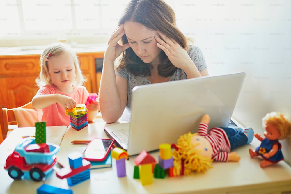 Exhausted and stressed mother working from home with toddler. Quarantine and closed daycare centres during coronavirus outbreak. Child making noise and disturbing woman at work. Freelance job with kid