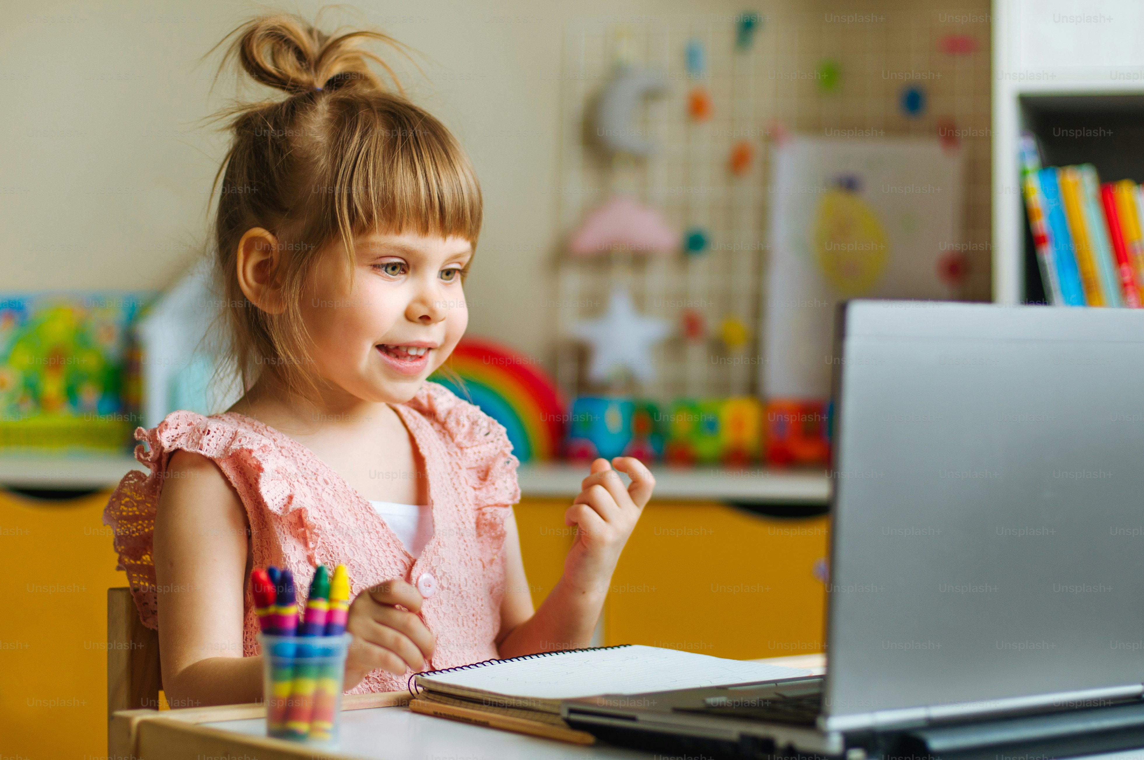 Happy little four years old girl watching online lesson using the notebook for distance learning sitting at the table in the kids room. Distance online learning concept.