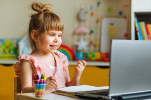 キッズルームのテーブルに座って、遠隔学習用のノートを使用してオンラインレッスンを見ている幸せな小さな4歳の女の子。遠隔オンライン学習のコンセプト。