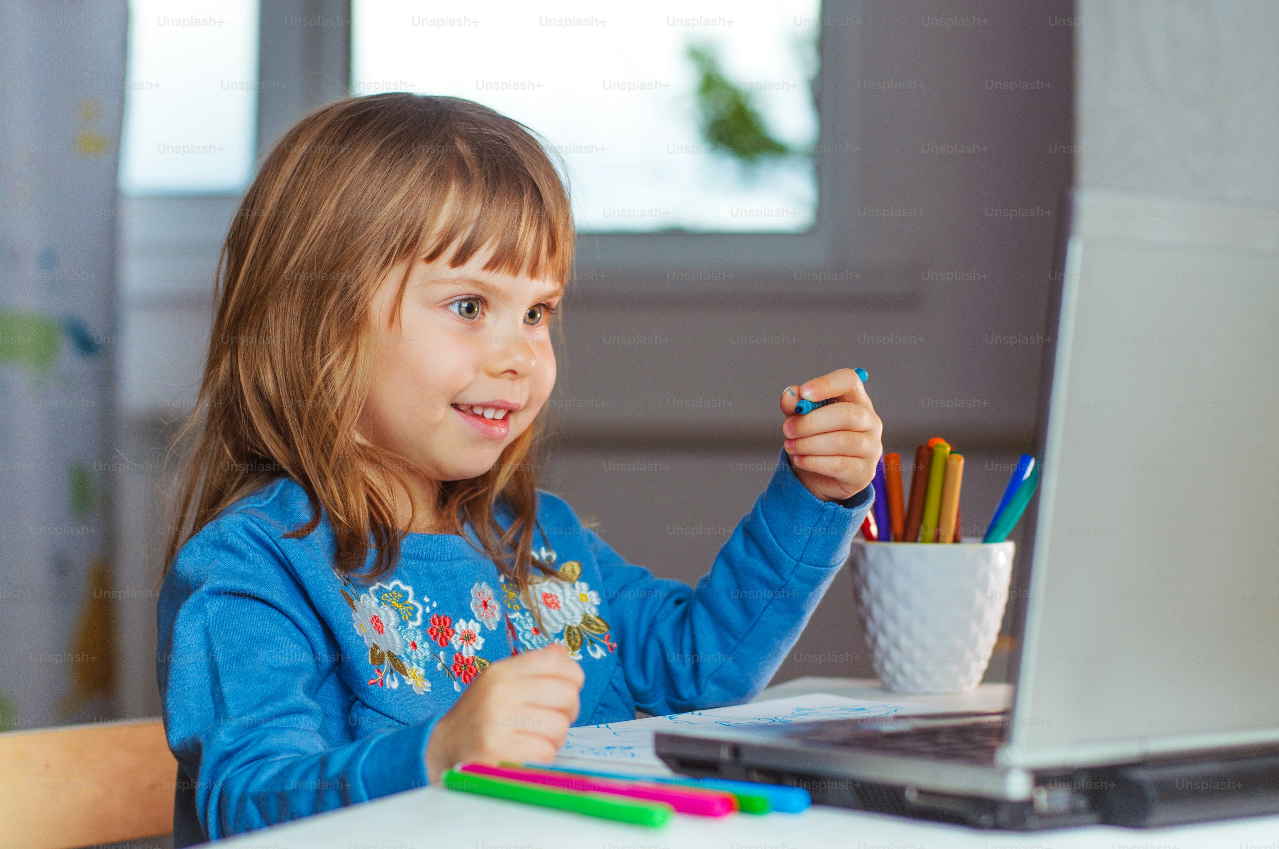 Happy little four years old girl doing homework using the notebook for distance learning sitting at the table in the kids room. Distance online learning concept.