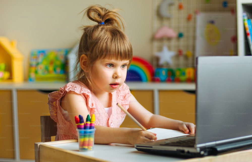 Menina estudante da escola primária desenhando ou escrevendo assistindo aula on-line usando o caderno sentado à mesa na sala das crianças. Conceito de ensino online à distância.