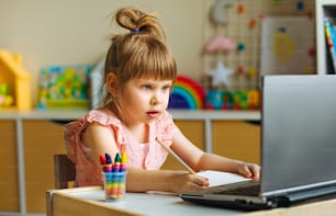 어린 소녀 초등학생이 어린이 방의 테이블에 앉아 노트북을 사용하여 온라인 수업을 보면서 그림을 그리거나 글을 씁니다. 원격 온라인 학습 개념입니다.