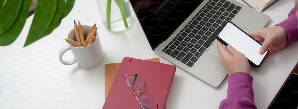 Cropped shot of young college girl using smartphone to find information for her thesis on white table with laptop and stationery