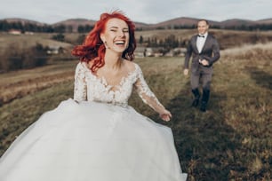 happy stylish bride and groom running and having fun in mountains at sunset light. gorgeous newlywed couple laughing, true feelings. emotional romantic moment. space for text