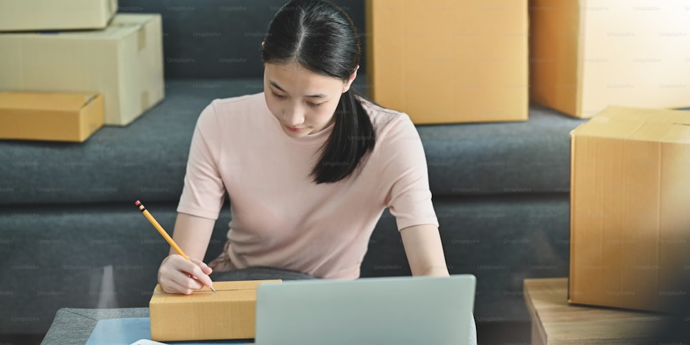 Photo of beautiful woman addressing/writing address on cardboard box while sitting in front her computer laptop over comfortable sitting room as background. Shopping online, SME, Entrepreneur concept.