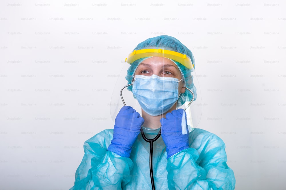 医師、看護師は、白い背景で隔離されたCovid-19コロナウイルスと戦うための保護スーツを着用する方法を示します。聴診器を装着したフェイスシールドを備えた完全防護服を着た医療従事者