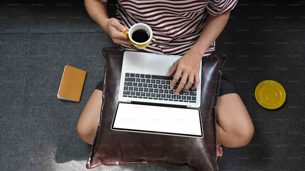 컴퓨터 노트북에 타이핑하는 동안 뜨거운 커피 컵을 들고 있는 창의적인 남자의 상위 뷰 이미지로, 무릎에 올려놓고 편안한 거실 위에 바닥에 앉아 배경으로 합니다.