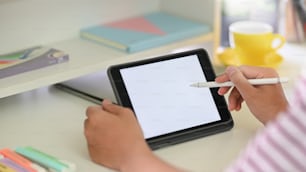 창의적인 사람들의 손이 흰색 빈 화면과 스타일러스 펜이 있는 자른 검은색 컴퓨터 태블릿을 들고 편안한 방 위의 현대적인 흰색 작업 책상에 배경으로 앉아 있는 자른 이미지.