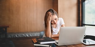 Femme stressée gardant la main sur la tête et regardant dans son ordinateur portable tout en souffrant d’un problème de travail et assise au bureau de travail en bois au-dessus d’un salon ordonné en arrière-plan.