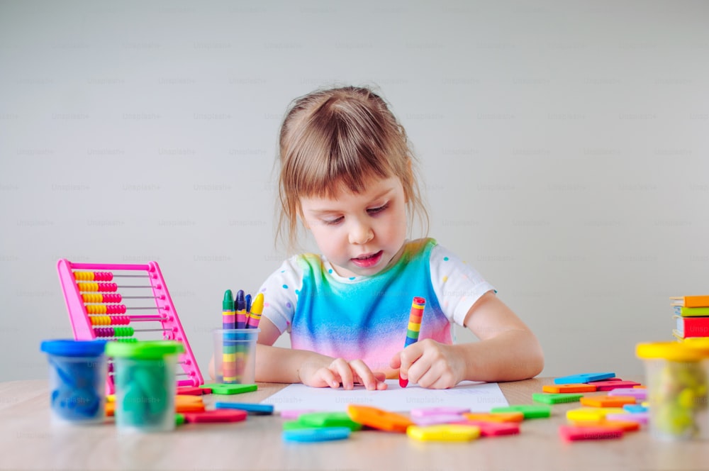 Beautiful little girl drawing with colorful wax crayons sitting at the table. Learning educational activities for children at home or in kindergarten.