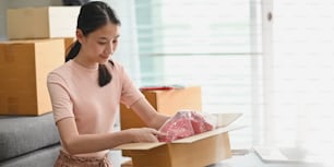 Photo of young entrepreneur woman packing her goods while sitting in front couch over comfortable sitting room as background. Shipping, Shopping online, Small business entrepreneur, SME, freelance.