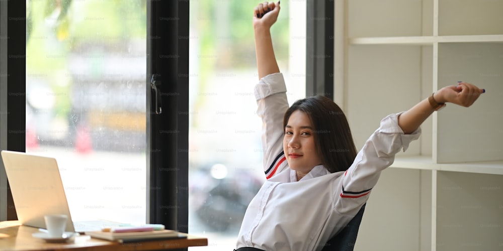Photo of beautiful woman working as secretary raised her hands up and stretching it while sitting and relaxing at wooden working desk over comfortable living room book shelf as background.