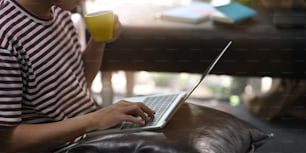 Cropped image of creative man drinking a coffee and using/typing on computer laptop that putting on his lap while sitting and relaxing at the leather couch over comfortable living room as background.