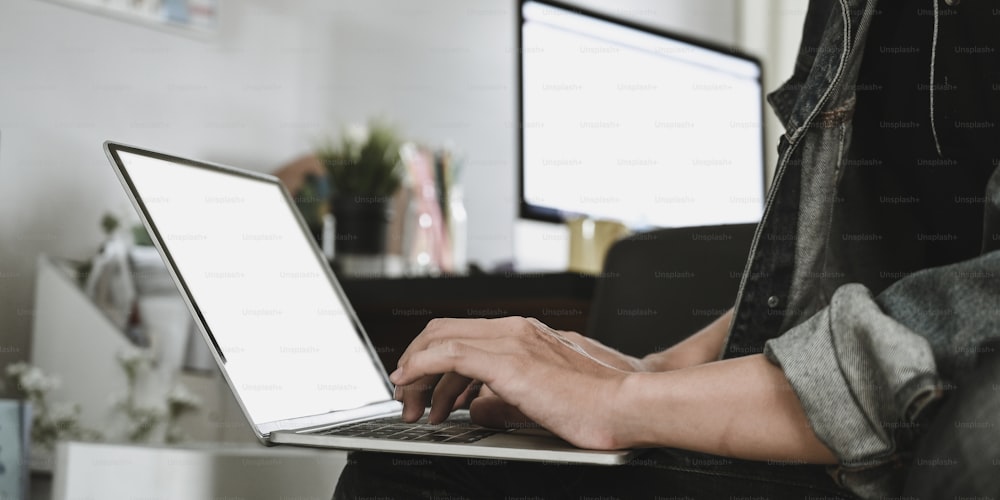 Image recadrée des mains d’un homme intelligent tapant sur un ordinateur portable à écran blanc blanc qui se met sur ses genoux au-dessus d’un salon confortable en arrière-plan. Concept de travail à domicile et de travail à distance.