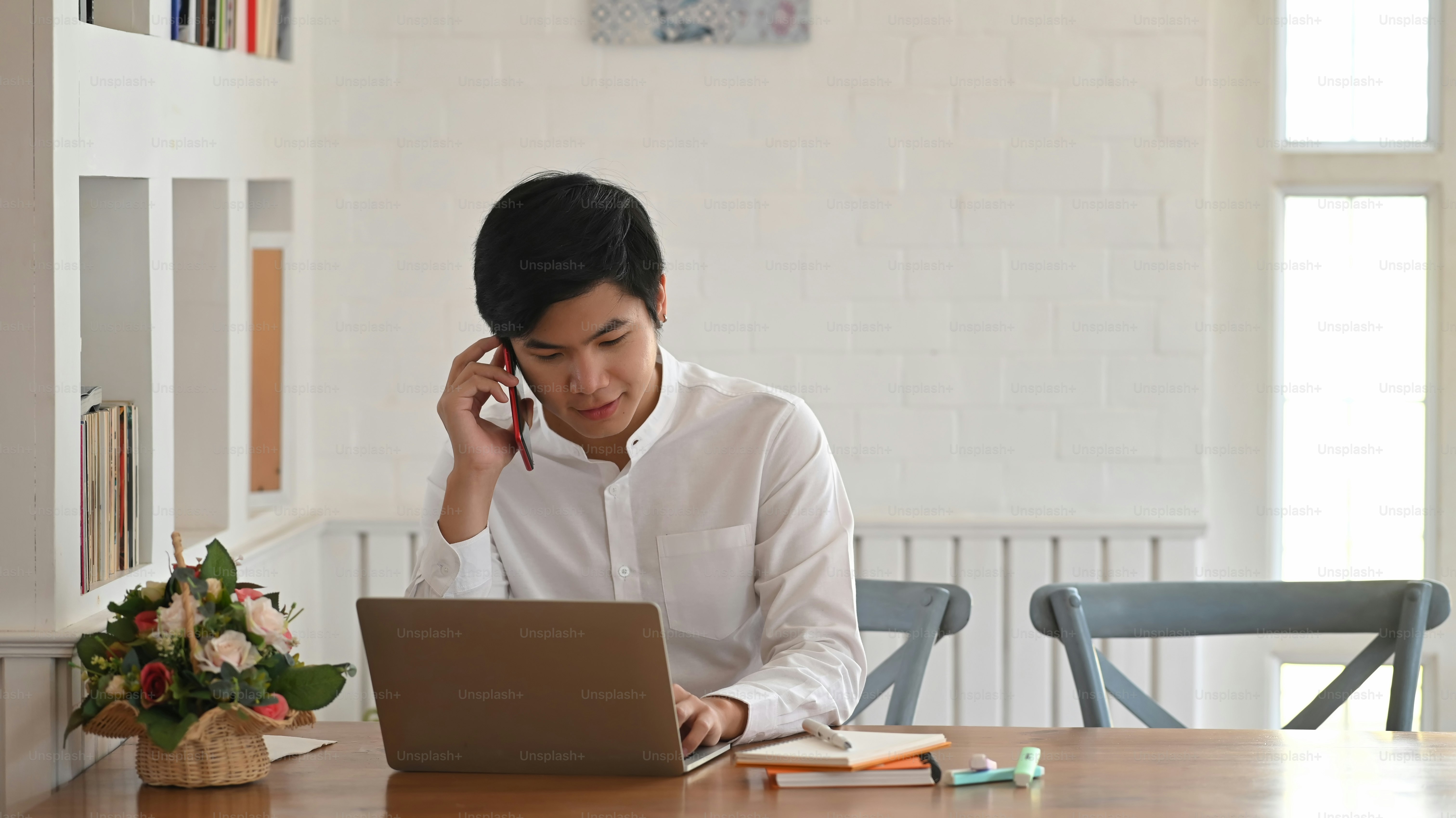 Foto do programador inteligente falando em seu telefone enquanto está  sentado e usando um laptop de computador na mesa de trabalho de madeira  sobre a confortável sala de estar como fundo. foto –, image size:3000x1687