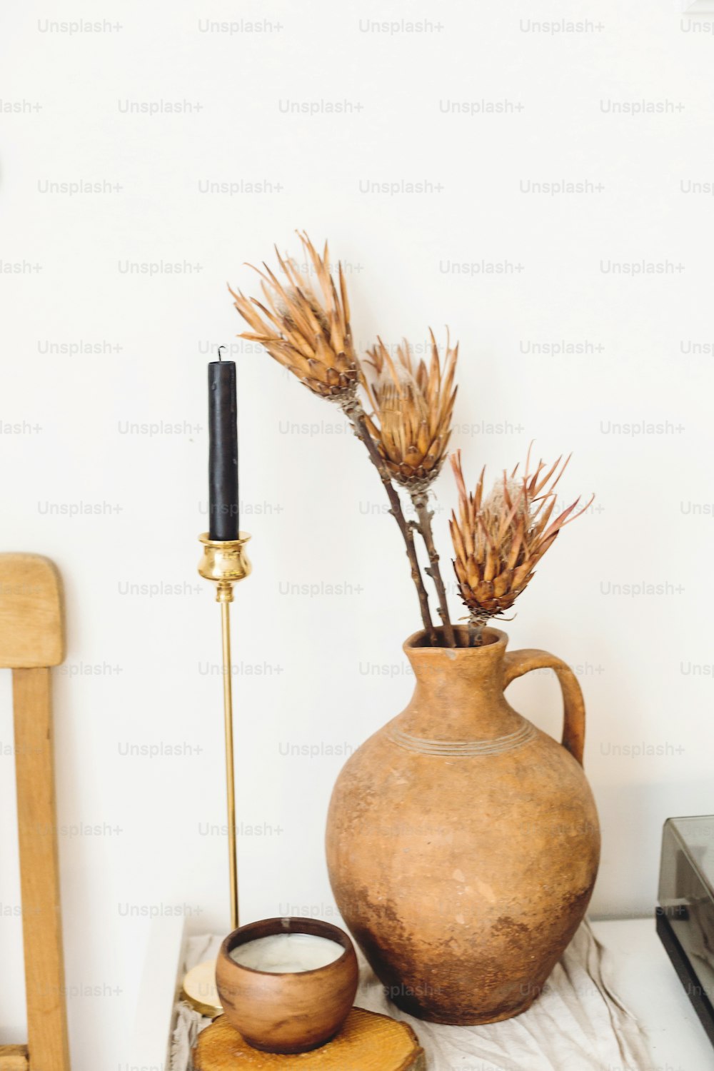 Dry protea flowers in old clay vase and candle on background of white wall in country home. Modern house decor. Stylish simple interior design elements. Rural house