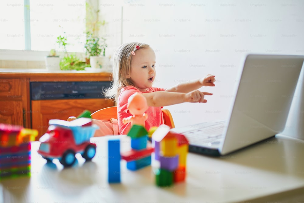 Happy toddler girl with laptop and toys. Kid using computer to communicate with friends, elderly relatives or kindergartners. Education or online communication for kids. Stay at home entertainment