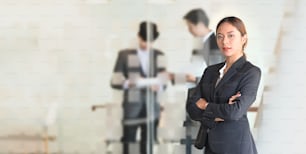 Mulher de confiança em terno formal preto em pé com continuar fazendo braço cruzado na sala de reuniões do escritório sobre seus colegas como pano de fundo.