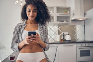 Contenta concentrata bella giovane donna dai capelli scuri in lingerie che invia un messaggio di testo sul suo smartphone