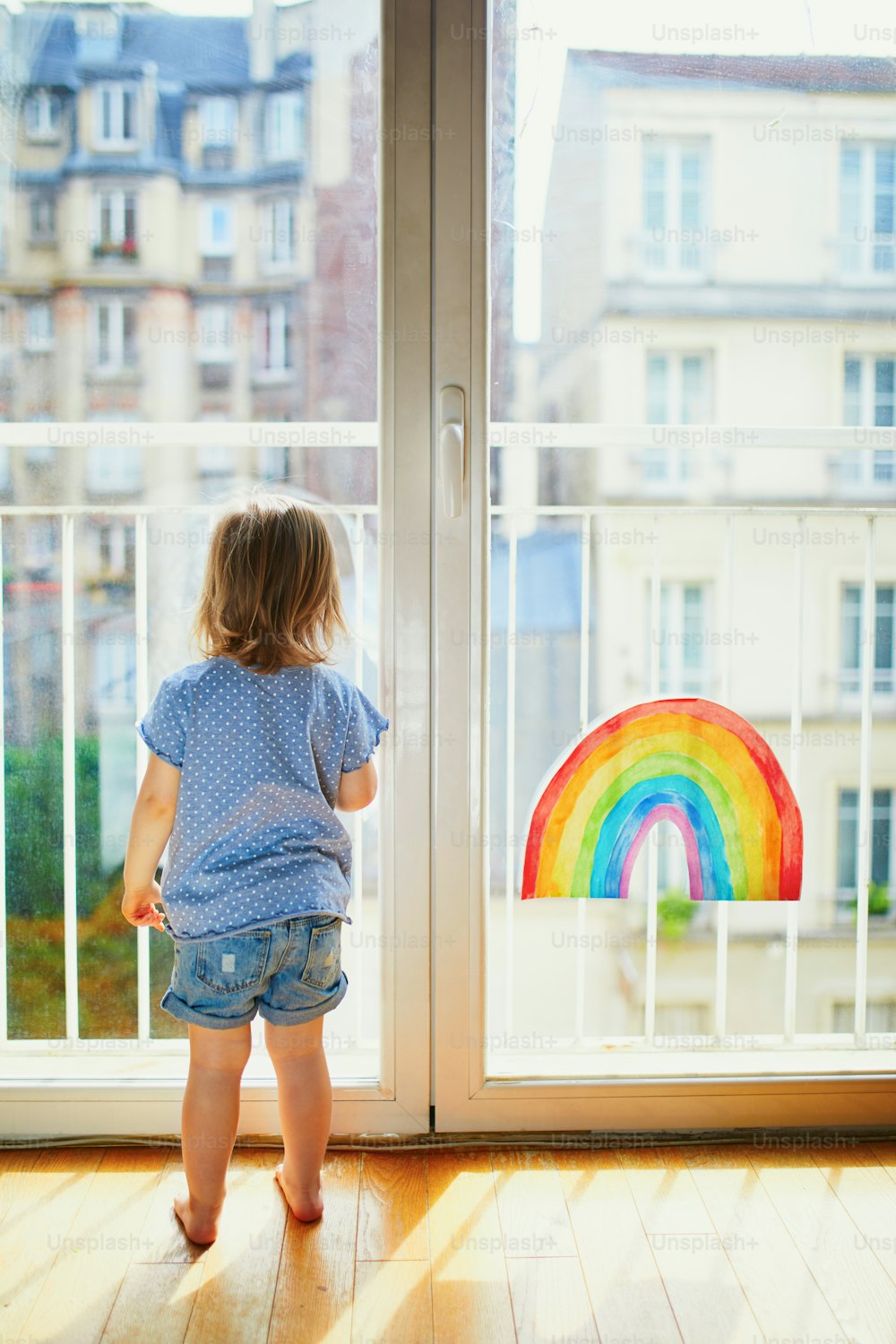 Adorable toddler girl looking through the window with rainbow drawing on it. Creative games for kids staying at home during lockdown. Self isolation and coronavirus quarantine concept