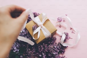 Concepto de feliz día de las madres. Caja de regalo que se abre a mano con cinta y flores lilas sobre papel rosa. Elegante tarjeta de felicitación floral. Ramo de flores lilas moradas con caja de regalo artesanal