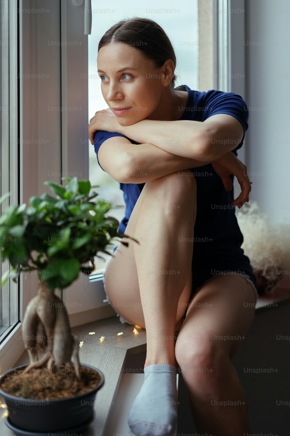 Rear view of beautiful woman looking out the window at the cityscape while staying home. Lady wears blue phantom T-shirt and shorts