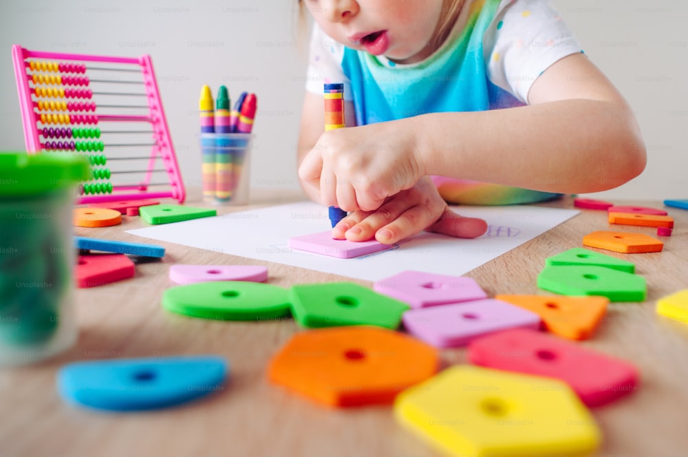 Primo piano della bambina che disegna intorno alla forma quadrata usando pastelli a cera colorati seduti al tavolo. Attività didattiche didattiche didattiche per bambini a casa o all'asilo.