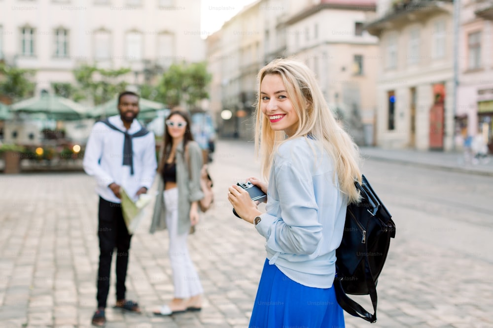 Outdoor summer smiling lifestyle portrait of pretty young blond woman having fun in the city in Europe with camera, and making travel photos for stylish multiethnic couple, African guy and white girl.