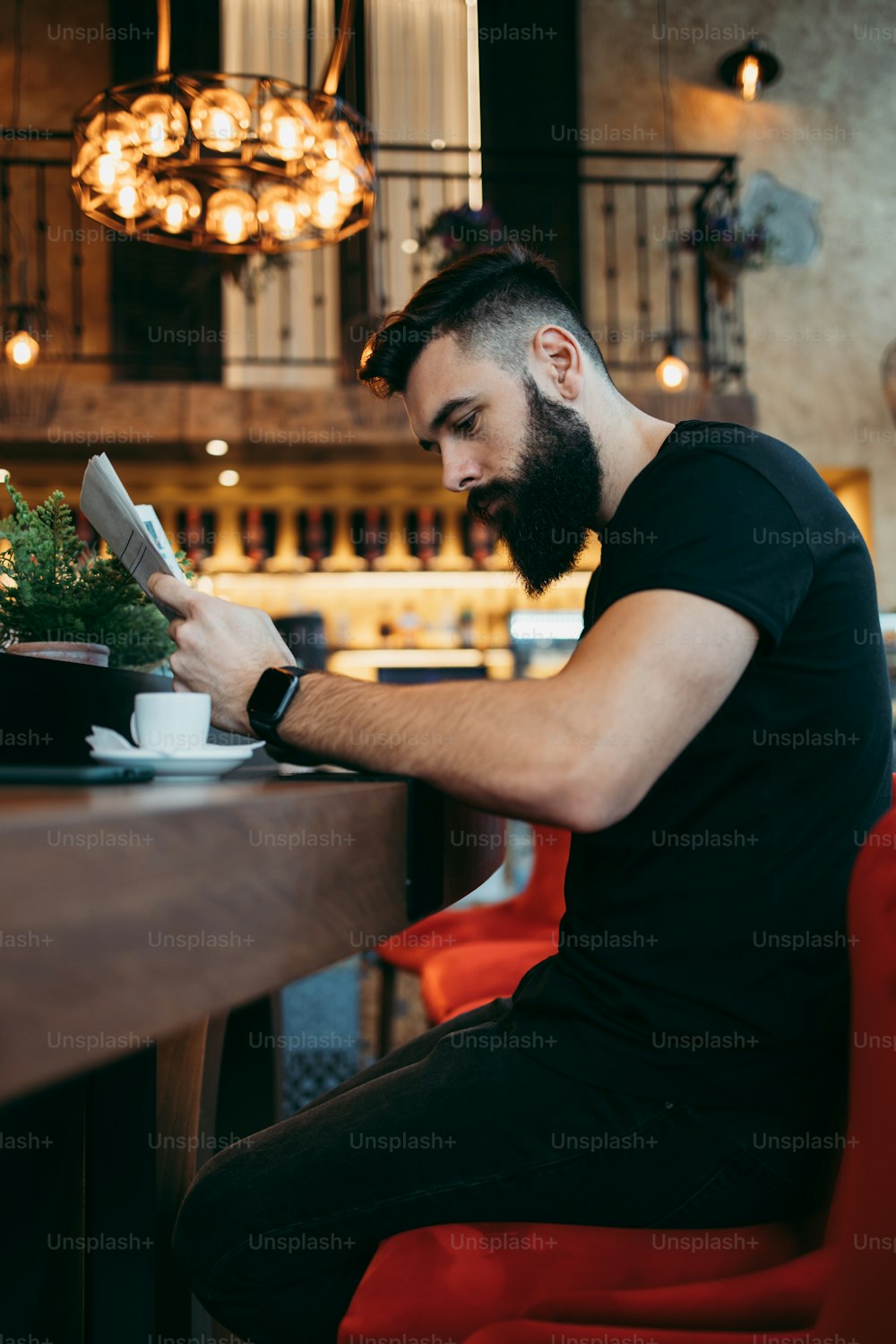 Hombre hipster adulto joven con una barba larga sentado en un café bar moderno, leyendo periódicos y reaccionando emocionalmente.