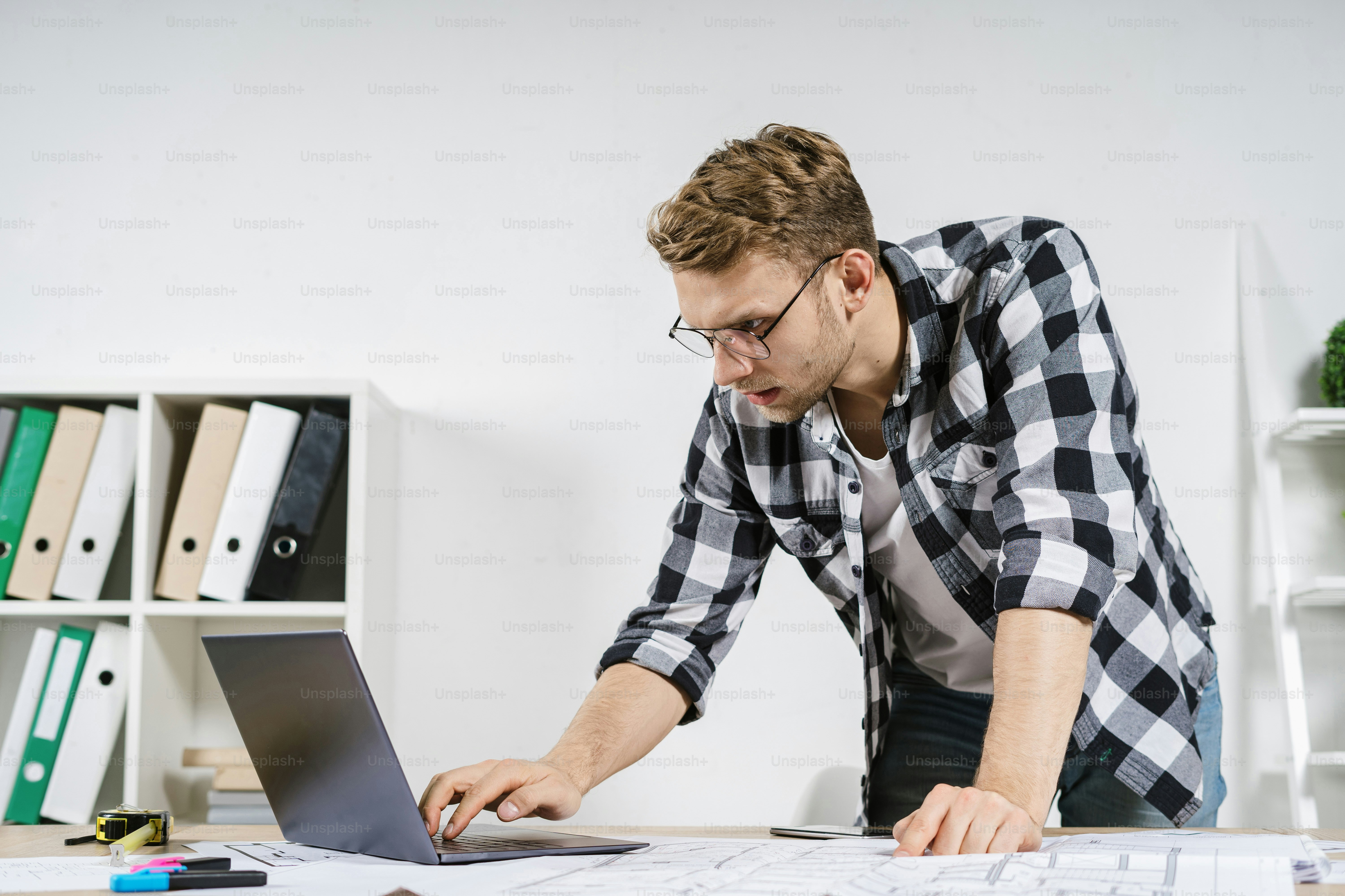 Focused and concentrated young adult architect working with blueprints in workspace office, using laptop computer, searching information online