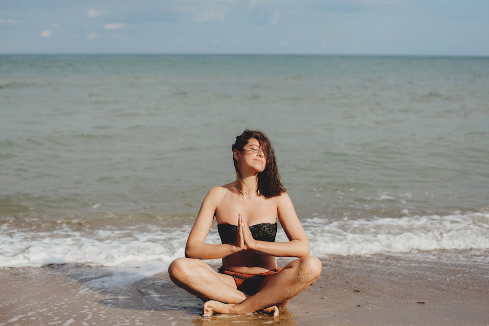 Giovane bella donna che pratica yoga sulla spiaggia, seduta sulla sabbia e medita. Concetto di salute mentale e cura di sé. Ragazza felice che si rilassa in riva al mare durante le vacanze estive