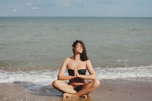 Young beautiful woman practicing yoga on the beach, sitting on sand and meditaning. Mental health and self care concept. Happy girl relaxing on seashore on summer vacation