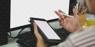 Cropped image of smart man holding a empty screen computer tablet and stylus pen while sitting in front of computer monitor with white blank screen over modern working room as background.