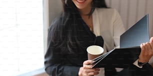 Imagen recortada de una elegante mujer de negocios sosteniendo una taza de café para llevar y un archivo de documentos mientras está sentada con un apoyo en las ventanas sobre una cómoda sala de estar como fondo.