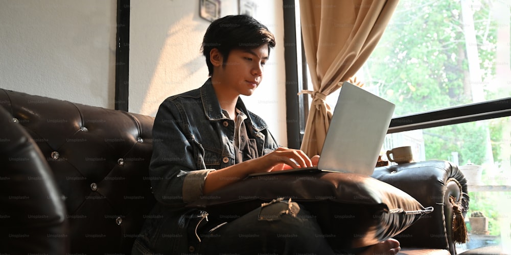 데님 셔츠를 입은 매력적인 남자가 컴퓨터 노트북에서 작업/타이핑을 하고 편안한 거실 위의 가죽 소파에 앉아 무릎을 꿇고 있는 사진.