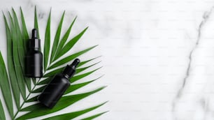 Black aromatic oil bottles on palm leaf on marble background. Hair serum lotion, herbal beauty products. Flat lay, top view.