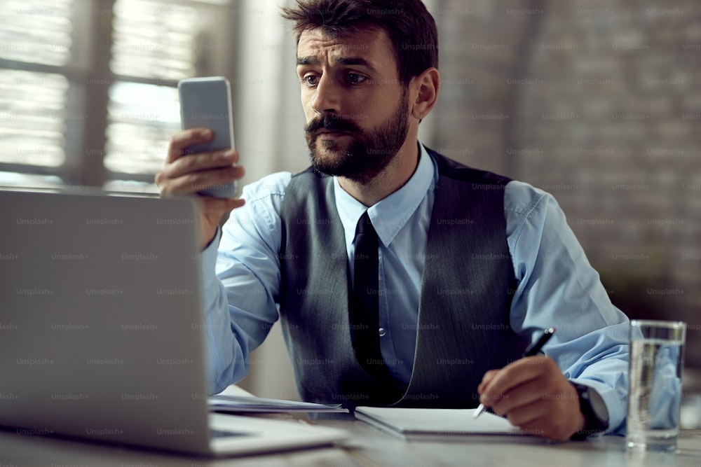 オフィスでメモを取りながら携帯電話とテキストメッセージを使用する男性起業家。