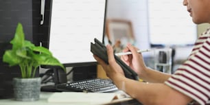 Imagen recortada de un hombre inteligente sosteniendo una tableta de computadora de pantalla vacía y un lápiz óptico mientras está sentado frente al monitor de la computadora con una pantalla blanca en blanco sobre una sala de trabajo moderna como fondo.