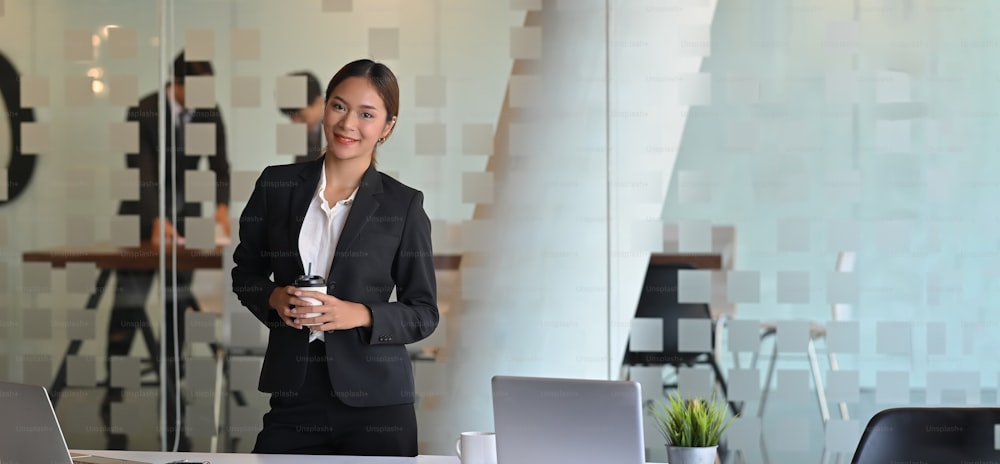 Confidence woman in black formal suit standing with holding a take-away coffee cup at the office meeting room over her colleagues.