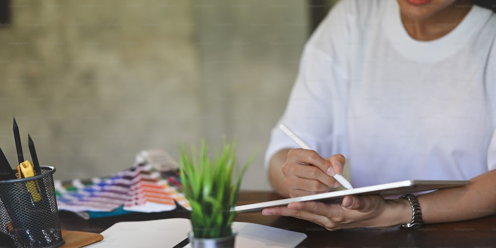 Imagem recortada de mulher criativa em camiseta branca segurando e desenhando no tablet do computador usando uma caneta stylus enquanto estava sentada na mesa de trabalho de madeira cercada por equipamentos de escritório.