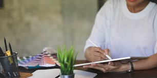 Imagen recortada de una mujer creativa en camiseta blanca sosteniendo y dibujando en una tableta de computadora usando un lápiz óptico mientras está sentada en el escritorio de trabajo de madera que está rodeado de equipo de oficina.