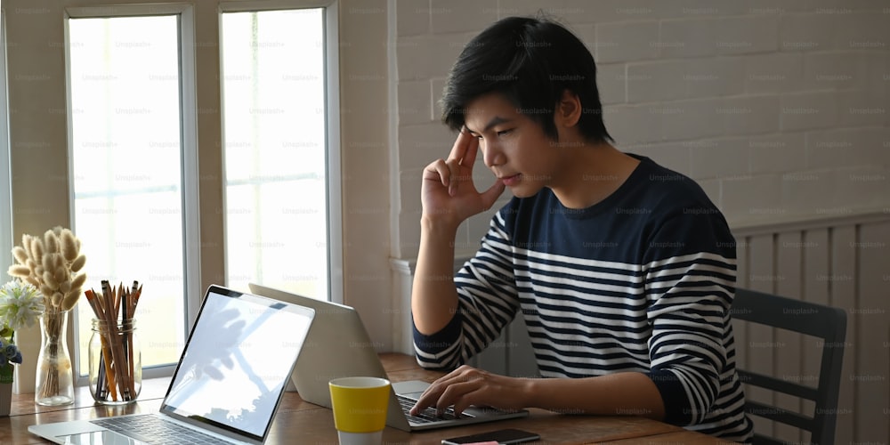 L'uomo creativo tiene la mano sulla testa mentre guarda nel computer portatile che mette sulla scrivania di legno e circondato da tazza di caffè, computer portatile.
